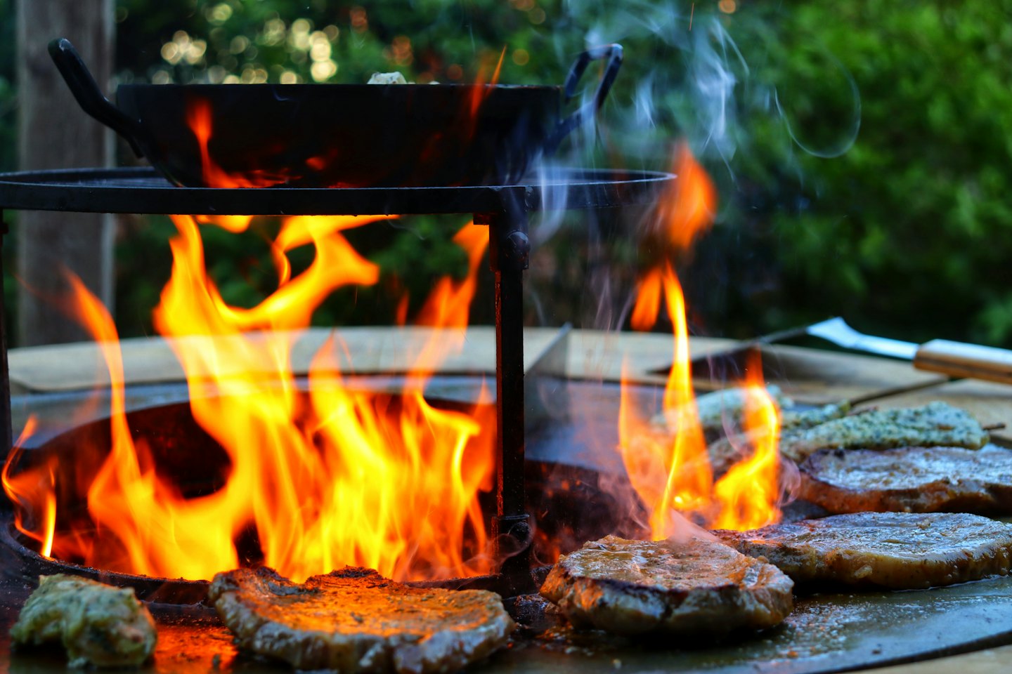 Steaks and skewers sizzle on an outdoor grill with orange flames.