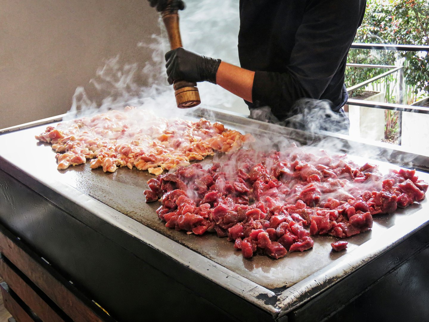 Chef seasons meat on a steaming flat-top grill.